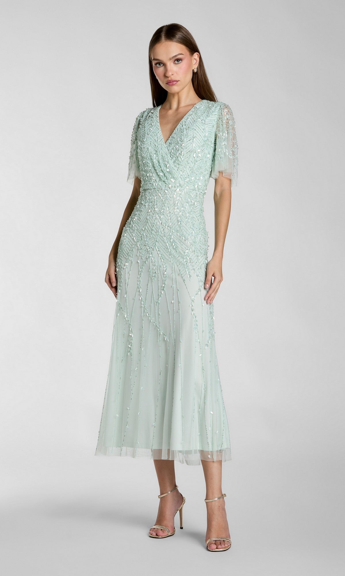 A woman stands in strappy heels against a plain background, wearing the Mac Duggal 35116 Beaded Midi Semi-Formal Dress in seafoam with short sleeves, a V-neckline, and a sheer overlay.